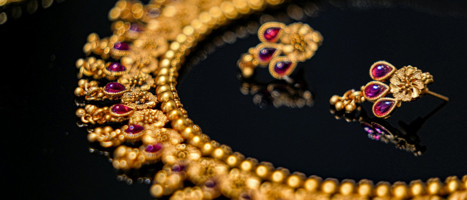 Ornate 22kt gold necklace with ruby-red accents and matching earrings on a dark background
